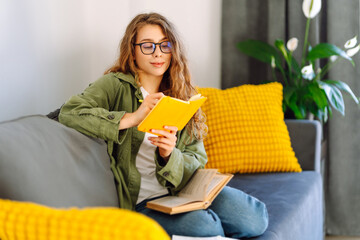 Portrait of smiling woman with yellow notebook in hands sitting on sofa in cozy room. Young female...