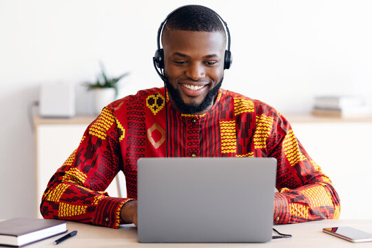 Distance Education. Smiling Black Man In African Shirt And Headset Watching Webinar On Laptop At Home, Having Online Lesson, Positive Millennial Guy In Ethnic Costume Sitting At Desk With Computer