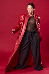 Fashionable asian woman in red leather coat and black pants posing confidently on a red background