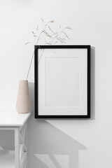 Minimalist black photo frame mockup on white wall beside a beige ceramic vase with delicate dried grass, styled in soft light and clean interior