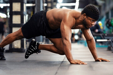 Plank work out concept. Active shirtless black man making work out on floor in modern male gym, side view. Motivated african american muscular guy bodybuilder planking knee to elbow, panorama