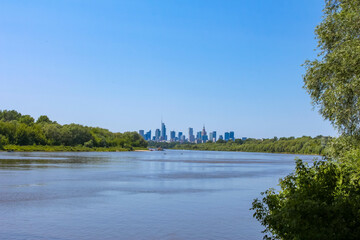 City skyline view across calm lake with green trees in foreground under clear blue sky on peaceful summer day