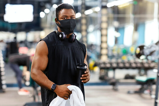 Sport and quarantine. African american bodybuilder in protective face mask holding water or protein and towel at gym, empty space. Black muscular man having break while exercising at modern gym