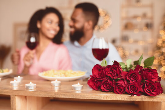 Fototapeta Romance And Bonding. Selective focus on bunch of red roses, bouquet of flowers lying on table. African american couple spending time together, having dinner, drinking wine in blurred background