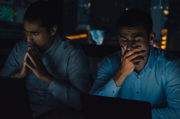 Two anxious professionals focus intently on their laptops in a dimly lit office during long working hours.