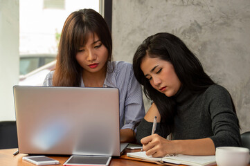 Two women collaborating on a project, one writing notes and the other working on a laptop in a modern workspace.