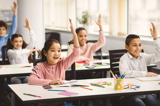 Academic Concept. Diverse group of multiethnic smart junior schoolchildren sitting at desks in classroom and raising hands for an answer, studying with pleasure. Positive young elementary students