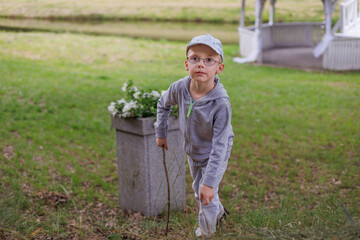 Curious boy in grey hoodie and cap plays with sticks on grass near a white gazebo by the pond,...