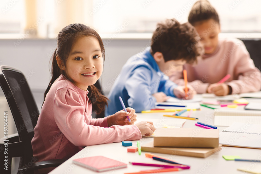 Poster academic concept. smiling junior asian school girl sitting at desk in classroom, writing in notebook