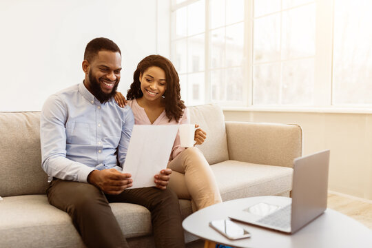 Portrait of cheerful african american couple sitting on the couch, man holding paper, working and reading letter with good news at home with his wife who is drinking coffee at home