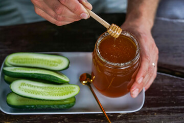 Person drizzling honey from a dipper into a jar, with fresh cucumber slices on a plate – natural food pairing and healthy snack concept.
