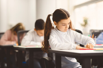 Portrait of small girl with ponytails sitting at table in classroom at primary school or kindergarten, writing or drawing in notebook. Reopening and return back to school after coronavirus quarantine