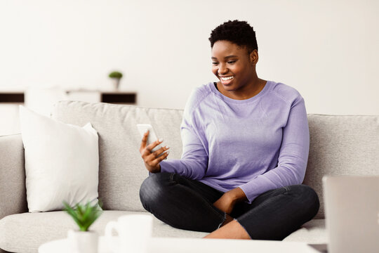 Positive young chubby black woman sitting on couch at living room, having video call on mobile phone, holding gadget, copy space. Happy african american lady having conversation on smartphone at home - Powered by Adobe