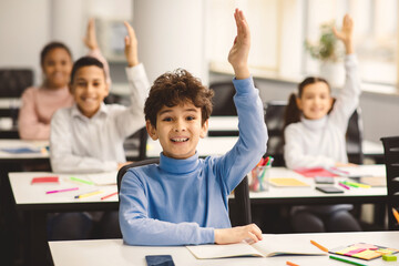 Education, Junior School, Learning and People Concept. Happy schoolboy raising hand for an answer, sitting at desk in classroom with diverse group of smiling classmates, studying with pleasure