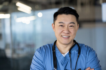 Close-up portrait of a young Asian male doctor standing in a blue uniform, arms crossed over his...