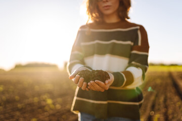 A young female agronomist holds fertile soil in a sunset field. A female farmer checks the quality...