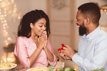 Marry Me. Smiling bearded african american guy holding and showing open box, offering marriage ring to his surprised excited fiance, getting engaged in restaurant or at home at romantic dinner