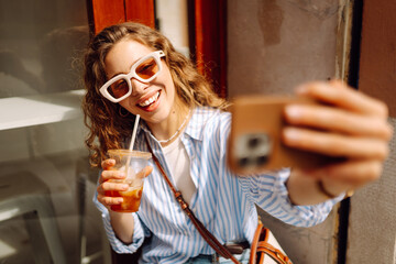 Portrait young woman drinks cooling drink and takes selfie on phone. Cheerful woman in glasses...