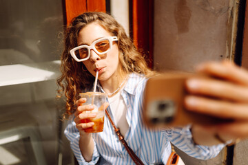 Portrait young woman drinks cooling drink and takes selfie on phone. Cheerful woman in glasses...