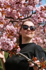Portrait of a confident woman in sunglasses standing among pink cherry blossoms in spring. Great for seasonal concepts, empowerment, lifestyle design, and outdoor visuals.