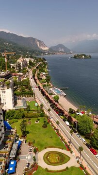 Drone video of charming towns on Lake Maggiore, Italy, including Stresa and Verbania, with historic grand hotels, sunlit roads, and vibrant lakeside life under bright midday sun