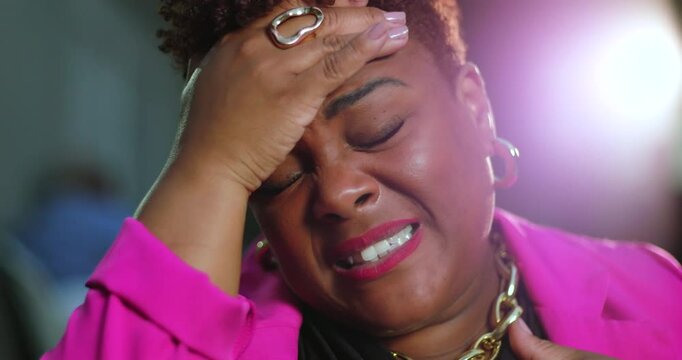 African American woman closing eyes and grimacing with hand on forehead, emotional expression of exhaustion and pressure in bright pink blazer and gold accessories