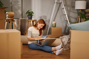 Woman is seated on the floor in a new home, surrounded by unpacked boxes while working on her...