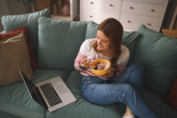 A woman casually sits on a couch, savoring dessert from a yellow bowl while engaging with her...