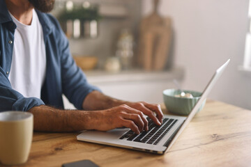 A man is typing on a laptop at a wooden table in a bright kitchen. A bowl of cereal sits beside him, and soft morning light fills the space, creating a warm atmosphere.
