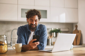 A man with curly hair and a beard enjoys his coffee while looking at his smartphone with a smile in a stylish kitchen during the morning hours.