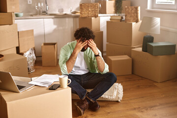 A person sits on a rug surrounded by numerous packed boxes, visibly stressed about the moving...