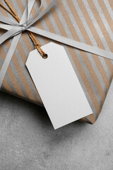 Close-up of a white paper tag tied with twine on a striped kraft gift box with a silver ribbon, placed on a concrete background, ideal for festive mockup use