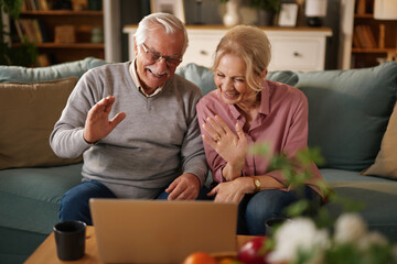 In a cozy living room, a smiling elderly couple sits on a sofa, waving and laughing while participating in a cheerful video call on their laptop, showing signs of connection and joy.