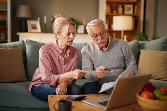 A Caucasian elderly couple sit on a blue sofa in their living room. The man holds a pen and papers while the woman points. A laptop, calculator, and bowl of fruit are nearby.
