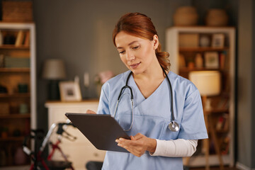 A young female Nurse wearing scrubs and a stethoscope looks down at a tablet device while visiting a Patient's home. There is a walker visible and bookcases in the background.
