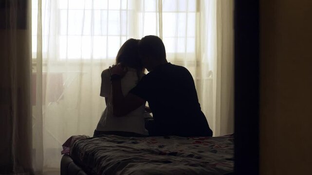 A couple sits together on a bed in a quiet room. The woman appears sad and emotionally exhausted. The man gently comforts her with a hug and kiss, offering support during a difficult time. 4K video
