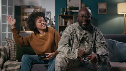 Medium full shot of excited African American male soldier in military uniform and 11-year-old polyethnic son playing console video game together, boy winning, cheering and giving dad high five