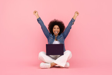Joyful young black woman sitting on floor on pink background with laptop on her lap, raising both arms in excitement, cheerful african american female celebrating success or online win, copy space