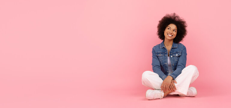 Young Black woman sitting cross legged on floor, looking up with a thoughtful and joyful expression, dreamy african american female posing on pink studio background, panorama with copy space