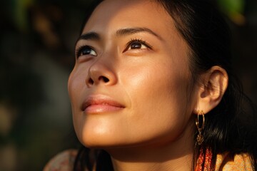Young woman gazing upward with serene expression, sunlight illuminating her face, casting gentle shadows, wearing gold earrings, dark hair pulled back, unfocused natural background