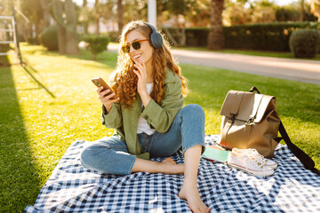 Young woman in casual clothes, wearing wireless headphones and using a phone, sitting on a mat on...