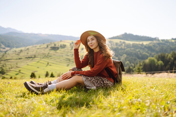 Young woman in hat and with backpack enjoying sunny weather on hillside. Beautiful female hiker feeling freedom on hiking mountain trail at sunset. Relaxation and nature concept. © maxbelchenko