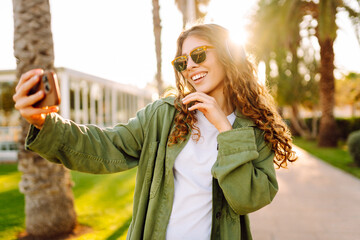 Happy woman with sunglasses and wireless headphones walks along an avenue with palm trees with a...