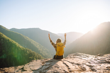 Beautiful woman hiker feels freedom standing on rocky ledge enjoying sunset in mountains. Young female hiker looks at mountain landscape on cliff. Hiking, adventure and nature concept.