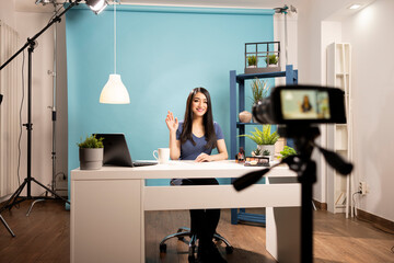 Beauty influencer seated at desk in studio with blue background, talking to online audience during...