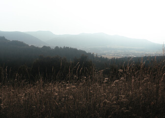 A wide mountain landscape with foggy peaks in the distance and dry, brown grasses in the foreground, showing summer haze