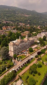 Drone video of Stresa town on Lake Maggiore, Italy, featuring historic grand hotels, bright midday sun, and cars driving along roads near the lake. Vibrant lakeside scene