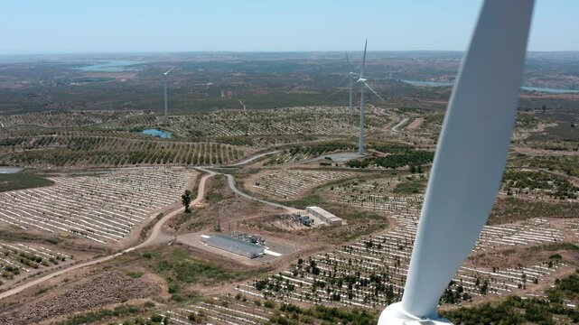 White wind turbine blades rotating, revealing expansive landscape with solar panels, verdant orchards, and serene water body against blue sky and distant horizon