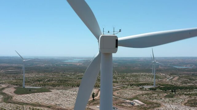 Large white wind turbine generating clean energy, rotating blades against a clear blue sky, overlooking an agricultural landscape with other turbines and a body of water in the background