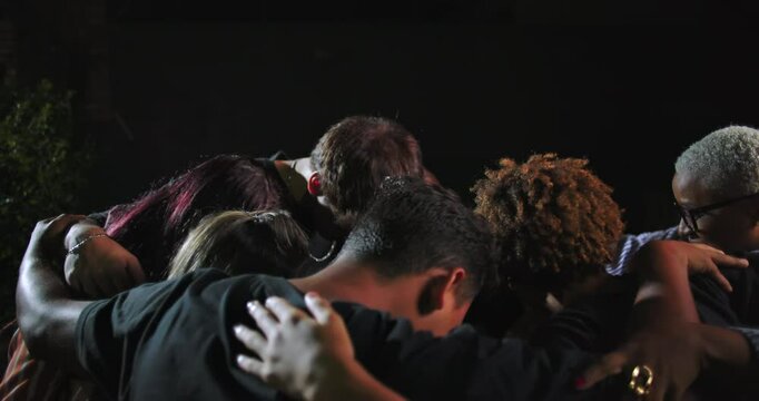 Group of people including African American and Latin Hispanic individuals embracing in tight circle, heads bowed in quiet nighttime prayer